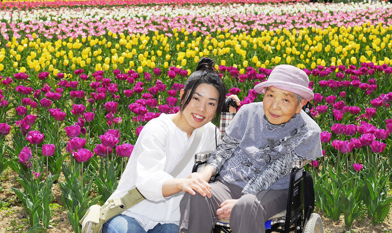 綺麗な花畑をバックに車いすに乗った女性高齢者とそばに寄り添ってしゃがむ女性